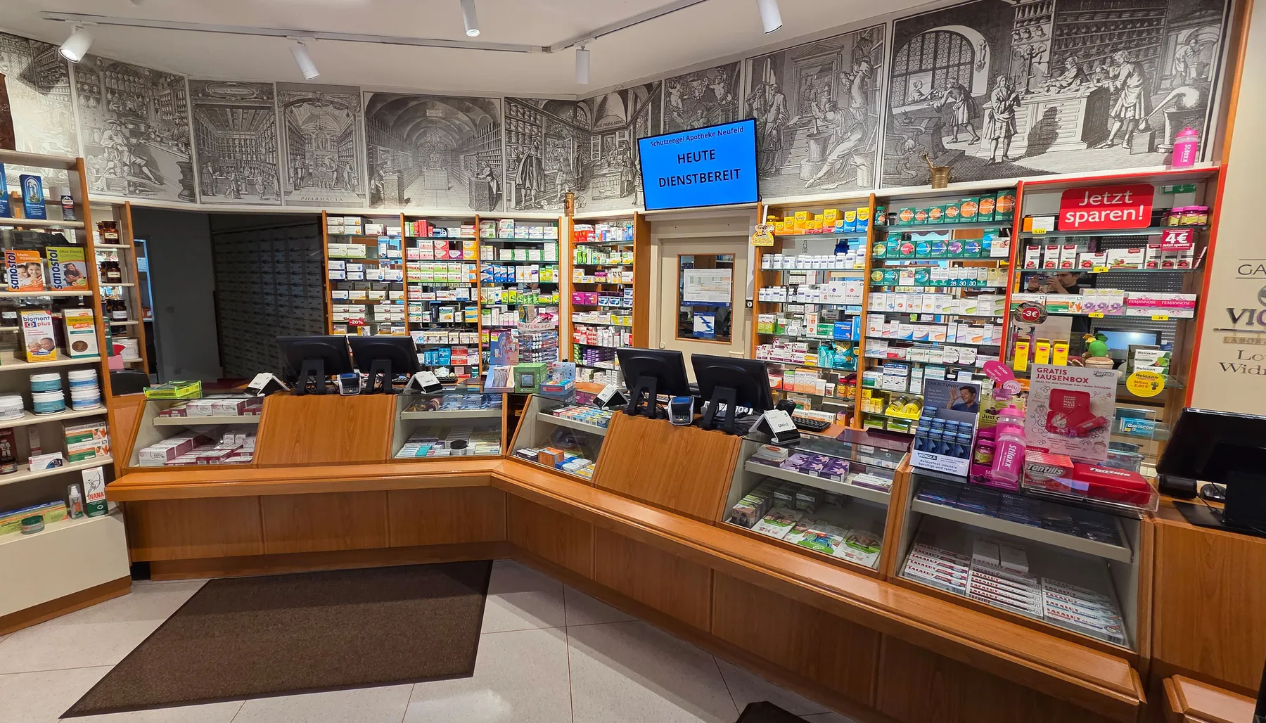 Blick in die Offizin der Schutzengel Apotheke: geschwungene Holztheke, Regale und Wandbilder historischer Apotheken